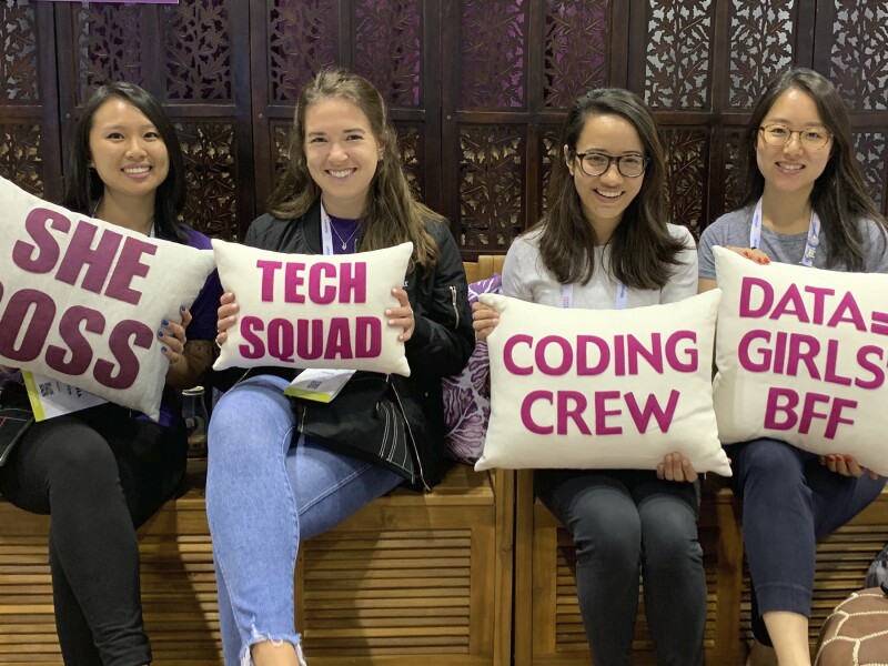 Four women sit smiling on a bench holding pillows with messages written on them, including "She Boss," "Tech Squad," "Coding Crew," and "Data = Girls' BFF".