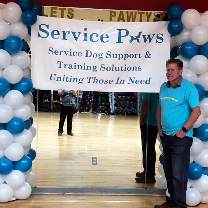 Man standing in front of balloon arch and banner that says "Service Paws, Service Dog Support & Training Solutions. Uniting those in need."