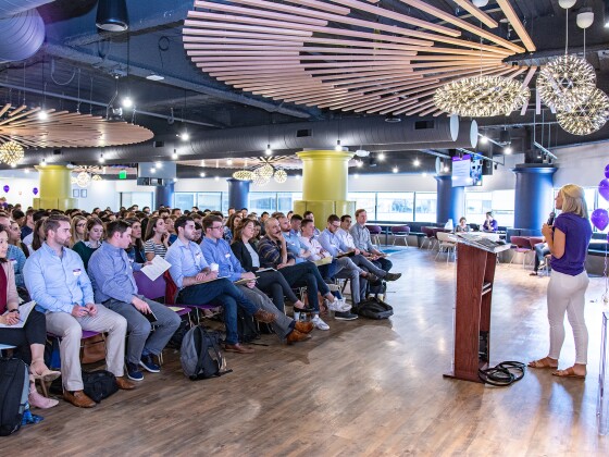 Image of crowd of people sitting down listening to a woman speak at a podium