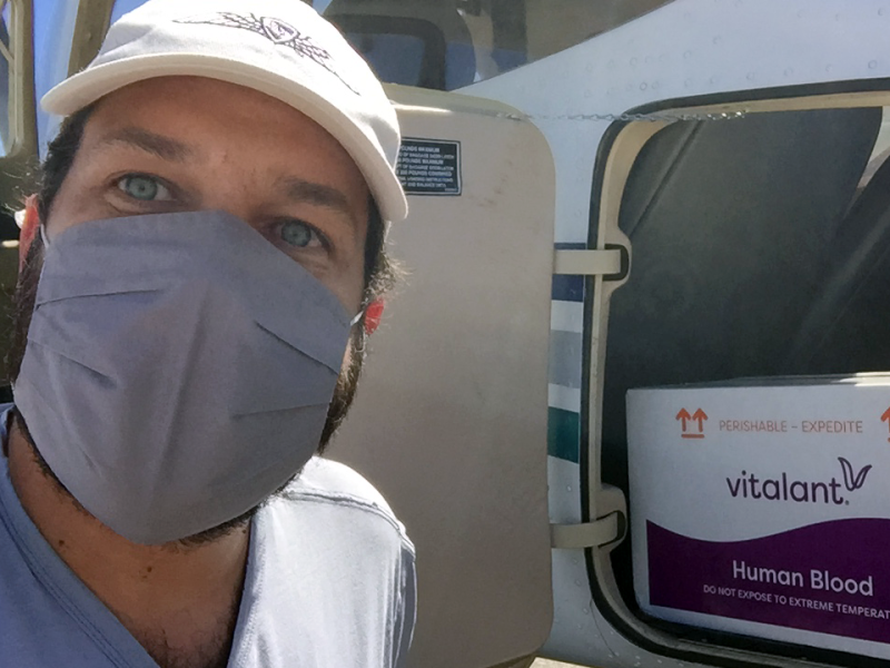 Man in a baseball cap standing in front of a plane loaded with a box labeled “Human Blood”.