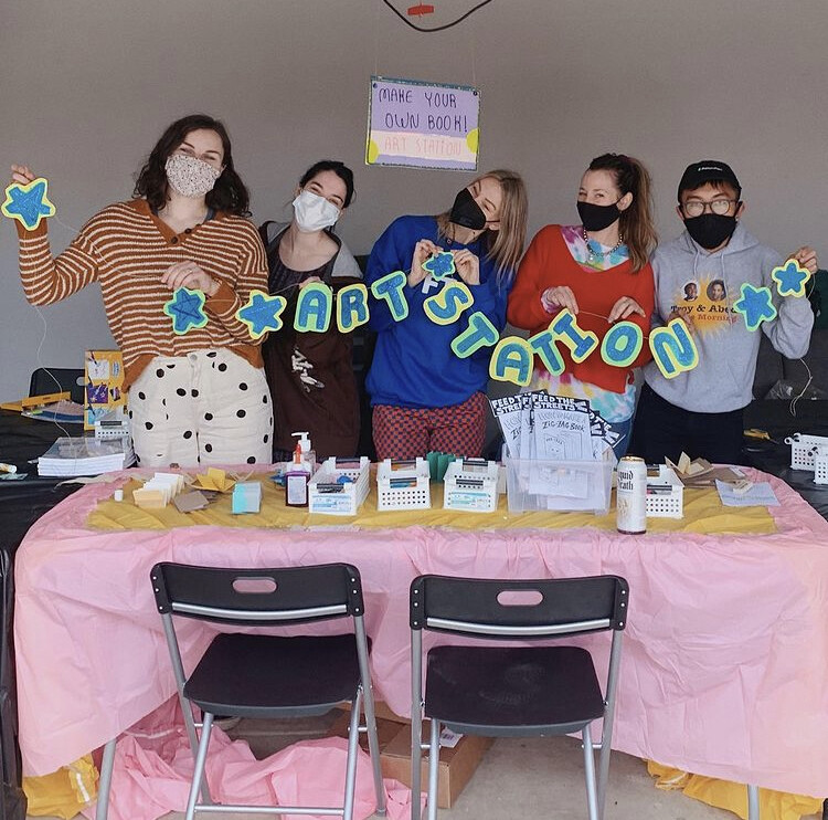 Four people holding a banner that says "Art station" in front of a table of arts and crafts.