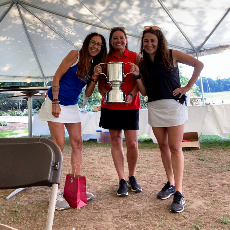 Three women in golfing outfits smiling and holding a large silver trophy.