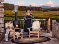 Kelly Clarkson stands on a wooden porch with log railing, leaning on her new furniture pieces (chairs). There's a beautiful Montana landscape stretching far behind her.