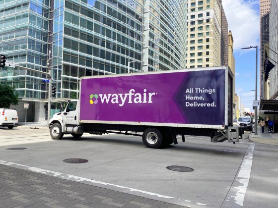 Image of Wayfair shipping truck on the road surrounded by tall city buildings providing e fulfillment service