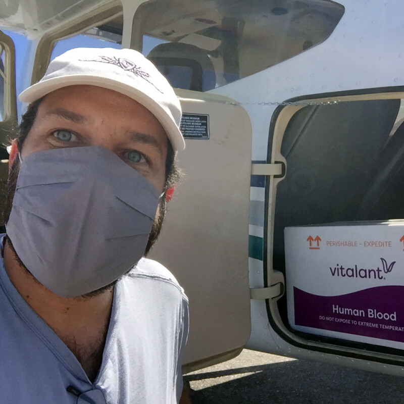 Man in a baseball cap standing in front of a plane loaded with a box labeled “Human Blood”.