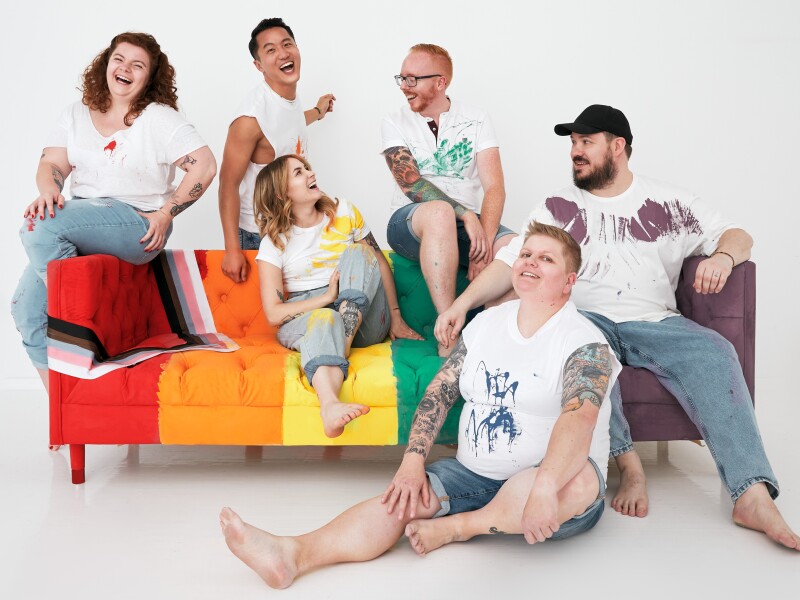 Six Wayfair employees sit on a pride-themed couch that they painted together, smiling and making eye contact with one another.