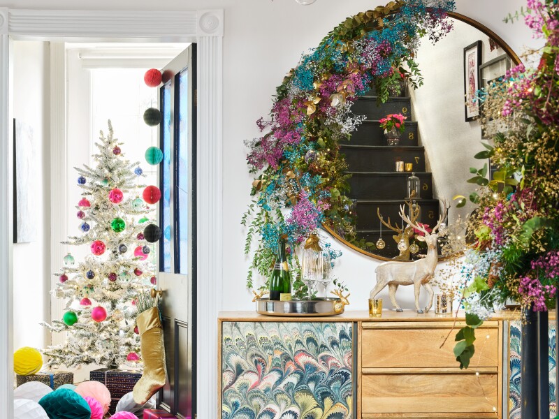 Holiday vignette with colorful faux silver tinsel tree, garlands on stairwell and mirror, and giant paper ornaments strewn across floor.